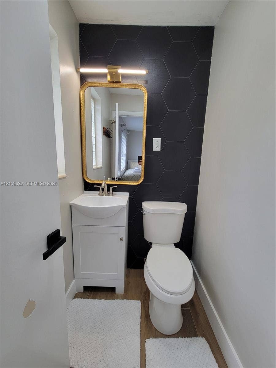 913 Lighthouse Drive, North Palm Beach, North Palm Beach, FL 33408 White pedestal sink with a metallic faucet, black hexagonal tile accent wall, gold-finish framed mirror, and wood-finish flooring