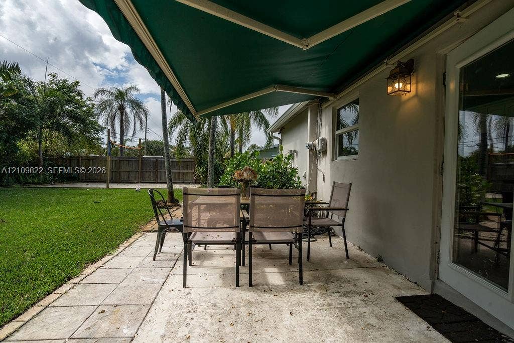 913 Lighthouse Drive, North Palm Beach, North Palm Beach, FL 33408 Backyard patio featuring concrete pavers, a retractable green awning, attached exterior wall-mounted lighting, and a fenced grass lawn