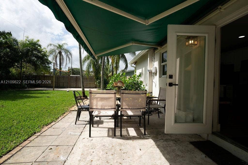 913 Lighthouse Drive, North Palm Beach, North Palm Beach, FL 33408 Paved stone patio featuring a retractable green awning, glass-paneled entry door, fenced perimeter, and mature palm landscaping