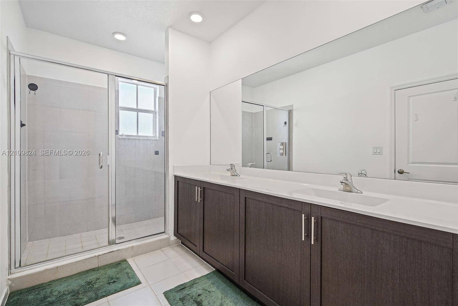 Double vanity with dark-finish cabinetry, solid-surface countertop, full-width wall mirror, and glass-enclosed shower with tiled surround