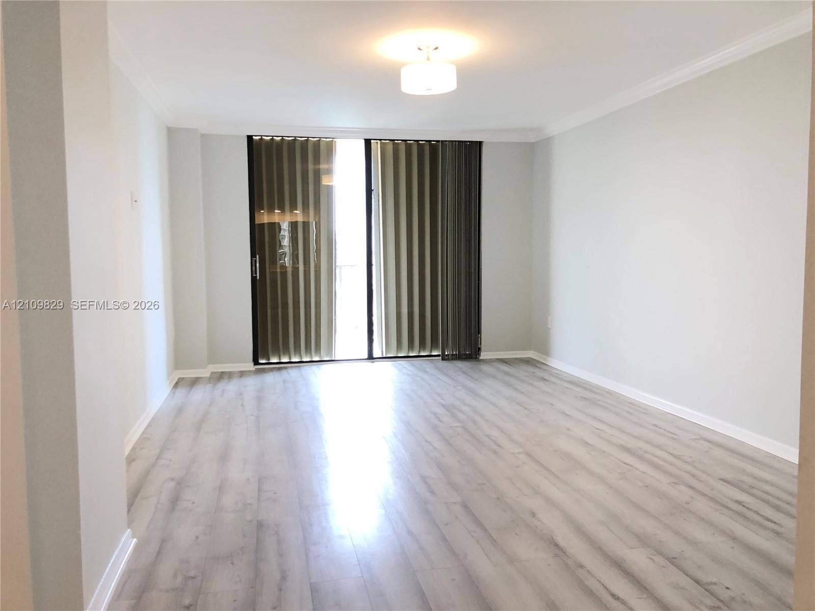 Living area featuring light-toned wood-finish flooring, crown molding, and sliding glass balcony doors with vertical blinds