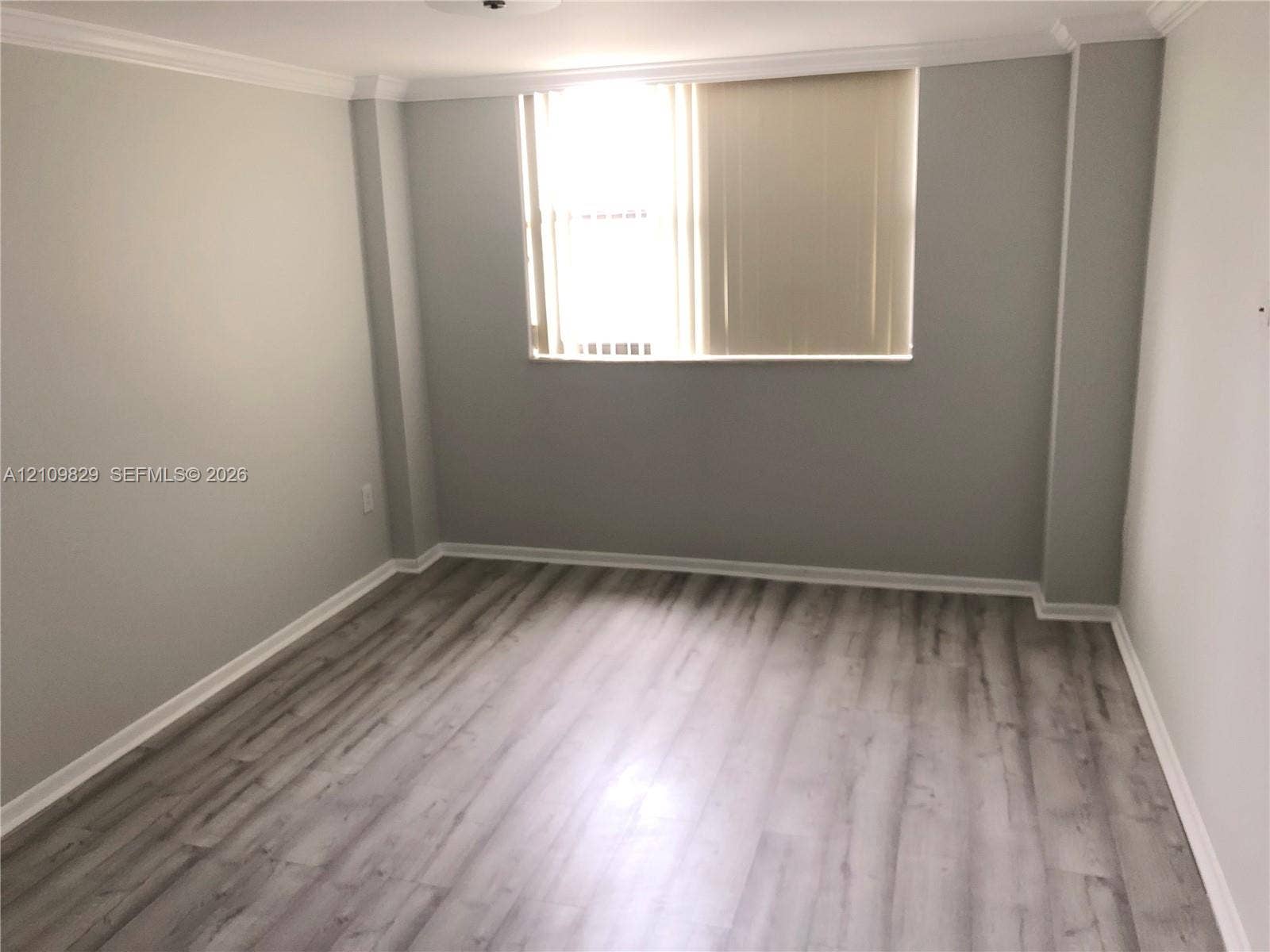 Neutral-toned interior featuring wood-finish flooring, crown molding, and a single window with vertical blinds