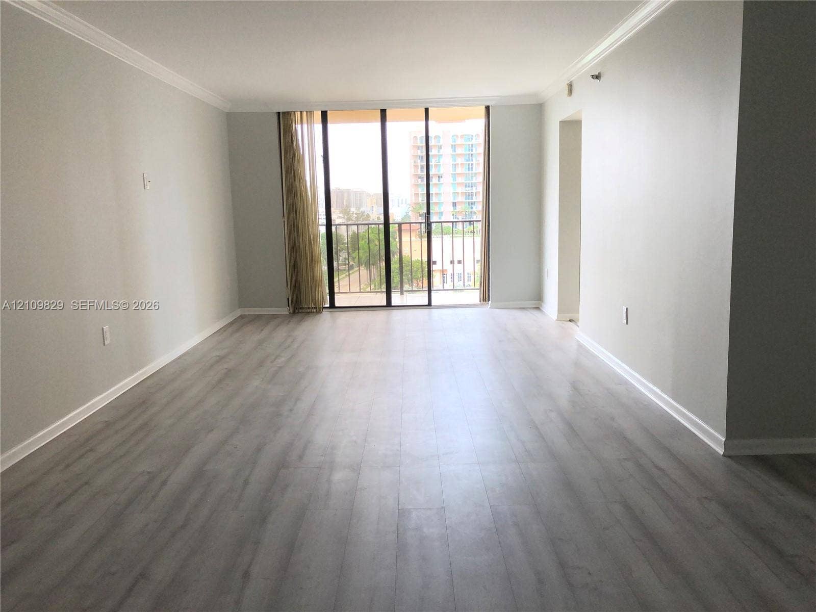 Open-concept room featuring light-toned wood-finish flooring, neutral wall paint, crown molding, and floor-to-ceiling glass sliding doors opening to a balcony