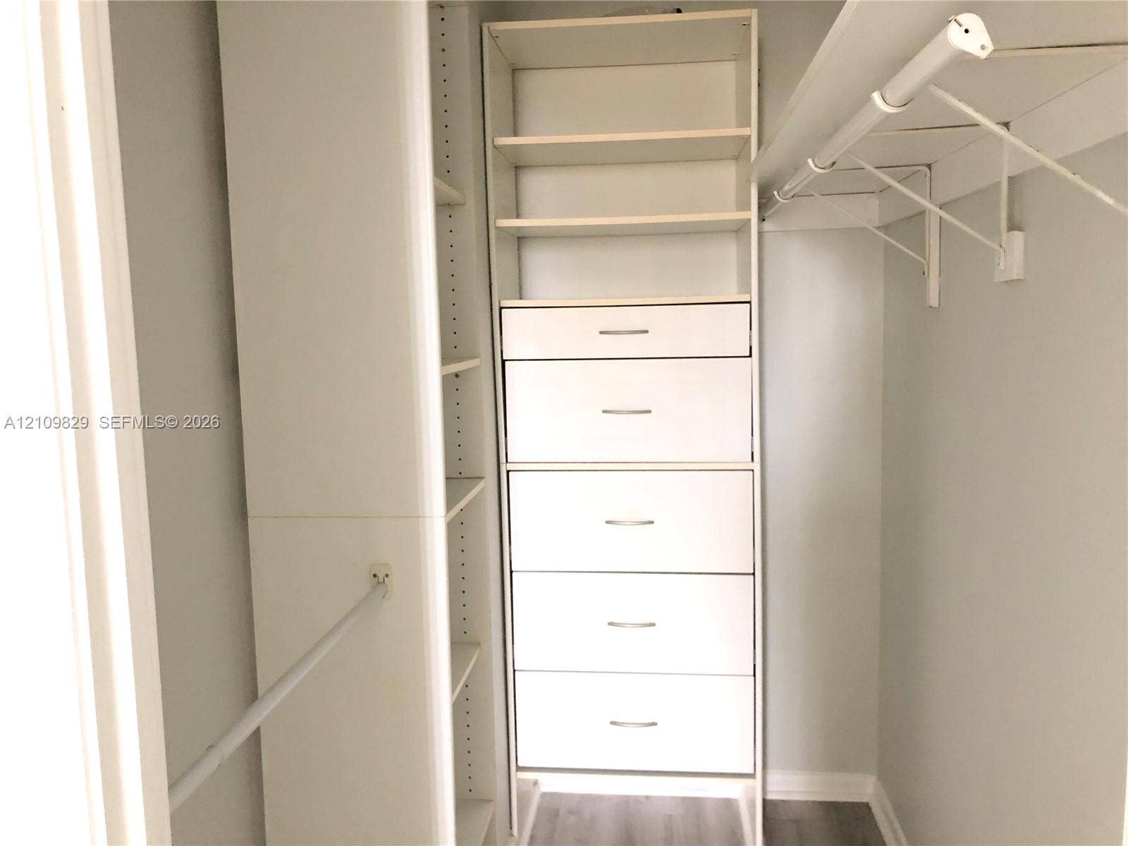 Walk-in closet featuring white built-in cabinetry, five integrated drawers, open shelving, and a wall-mounted garment rod over wood-finish flooring