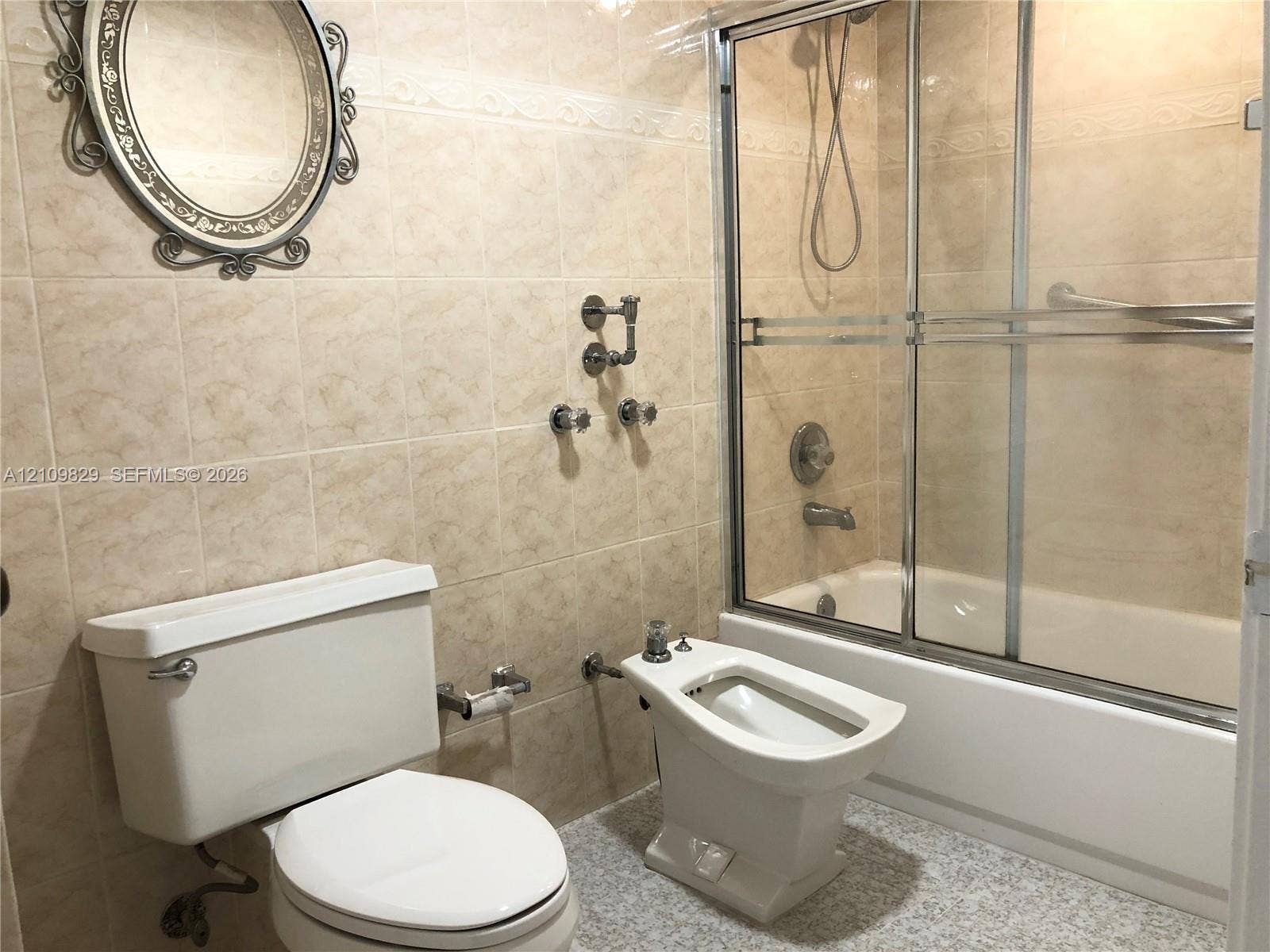 Bathroom featuring wall-to-wall ceramic tile, a soaking tub with a sliding glass enclosure, a pedestal bidet, and a wall-mounted metallic mirror