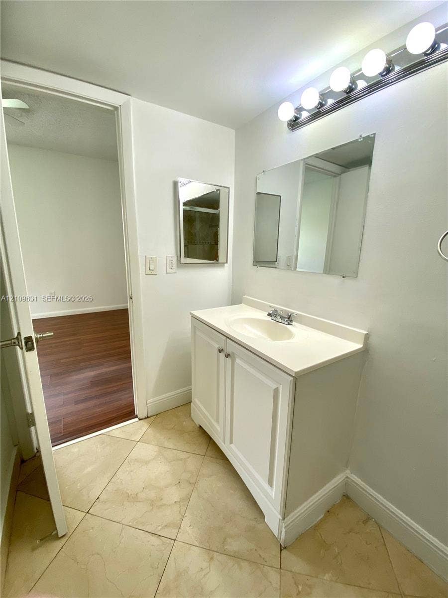 Bathroom vanity featuring a white cabinet base, integrated countertop and sink, wall-mounted multi-bulb lighting fixture, rectangular mirror, and neutral-tone tile flooring