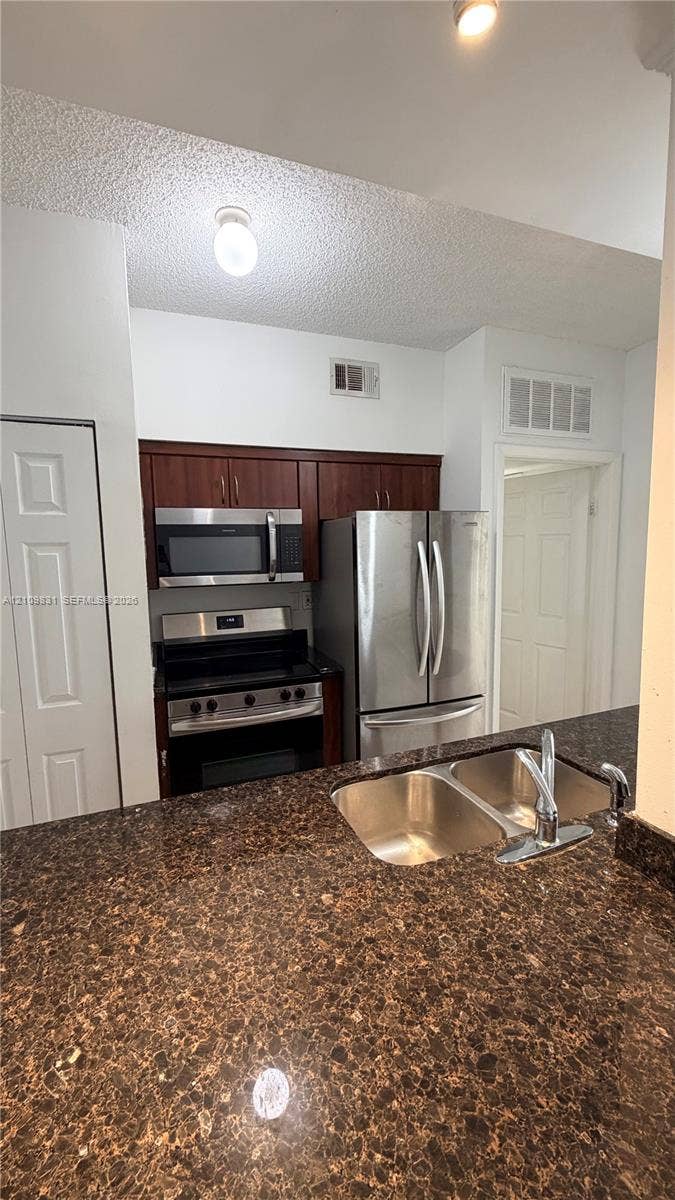 Kitchen featuring dark wood-finish cabinetry, stainless steel appliances, a multi-tone stone-finish countertop, and a double-basin stainless steel sink with a high-arc faucet