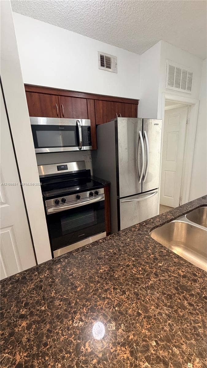 Kitchen featuring dark wood-finish cabinetry, stone-patterned countertops, a dual-basin stainless steel sink, and stainless steel appliances including a range, microwave, and refrigerator