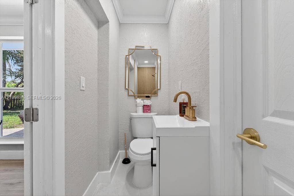 756 Cinnamon Road, North Palm Beach, North Palm Beach, FL 33408 Powder room featuring a white vanity cabinet, a gold-toned high-arc faucet, a geometric-framed wall mirror, and crown molding