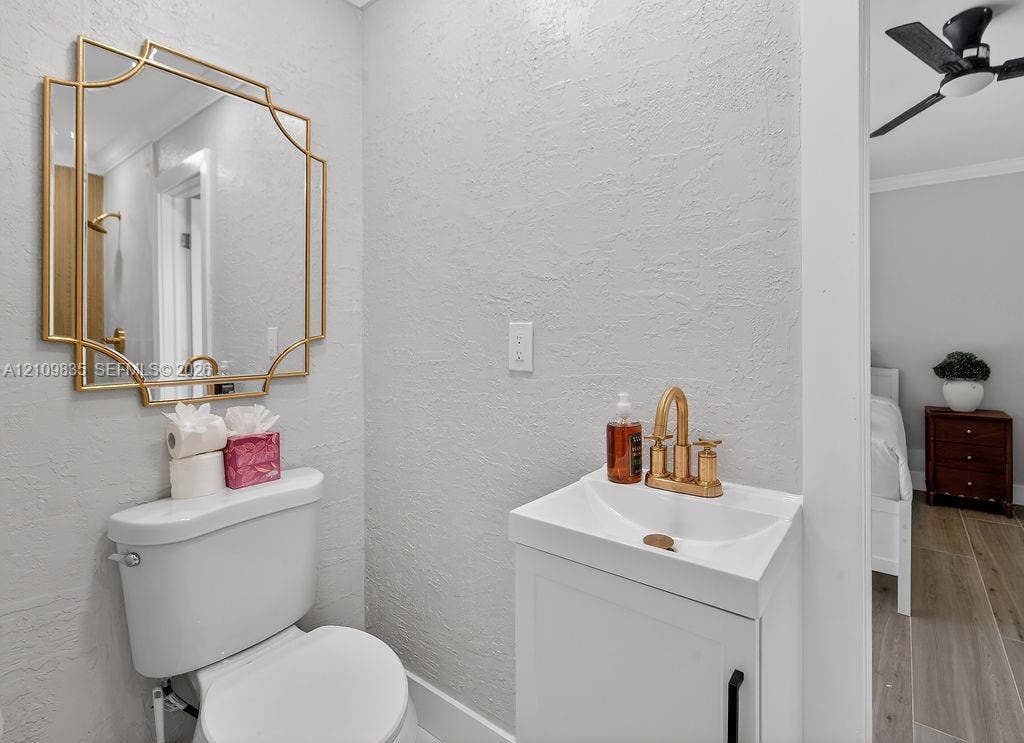756 Cinnamon Road, North Palm Beach, North Palm Beach, FL 33408 White vanity cabinet with an integrated sink basin, metallic gold faucet, geometric gold-framed mirror, and a ceiling-mounted fan