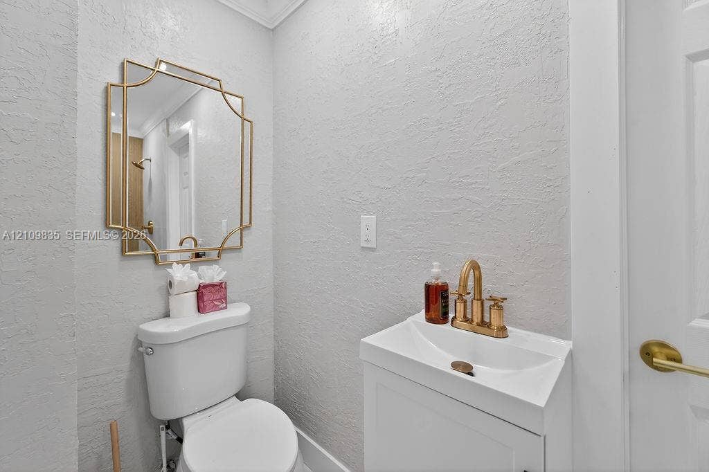 756 Cinnamon Road, North Palm Beach, North Palm Beach, FL 33408 Wall-mounted vanity featuring a white basin, brushed gold widespread faucet, geometric gold-framed wall mirror, and two-piece toilet