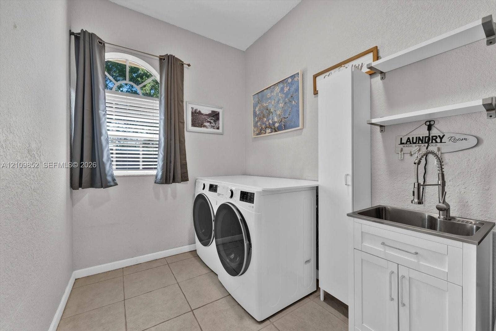 7901 SW 97th Street, Miami, FL 33156 Laundry room featuring ceramic tile flooring, an arched window, a white utility cabinet, and a stainless steel sink with a high-arch pull-down faucet and wall-mounted shelving
