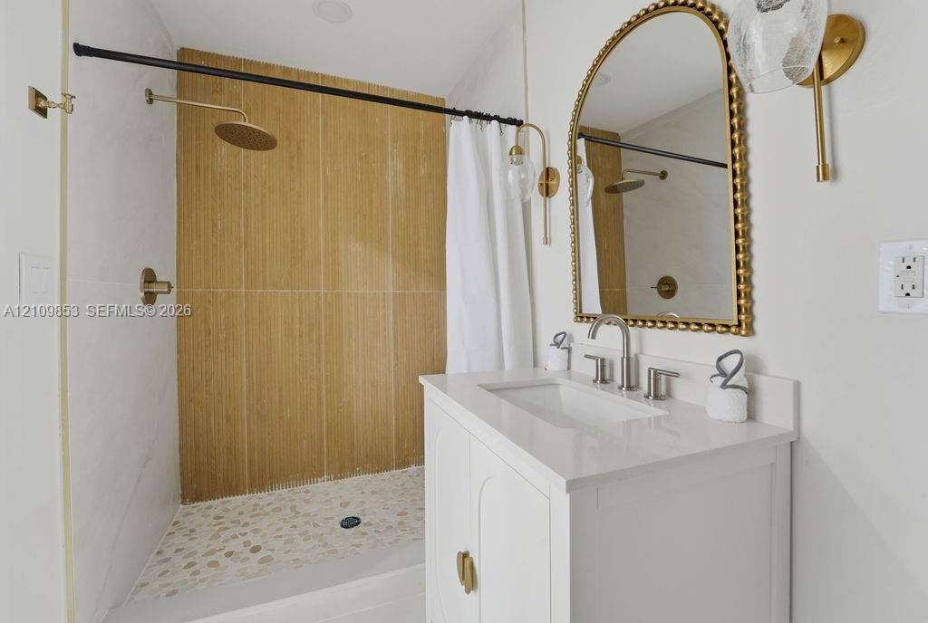 800 Alamanda Drive, North Palm Beach, North Palm Beach, FL 33408 Bathroom featuring a white vanity with a quartz countertop, arched beaded mirror, wall-mounted brass light fixtures, and a shower with wood-tone tile and river stone flooring