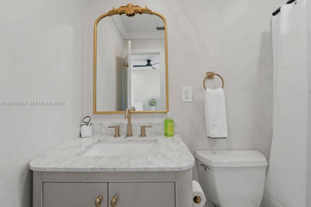 800 Alamanda Drive, North Palm Beach, North Palm Beach, FL 33408 Bathroom featuring a light-toned vanity with stone-surfaced countertop, under-mount sink, brass-finish widespread faucet, arched decorative wall mirror, and porcelain wall tile