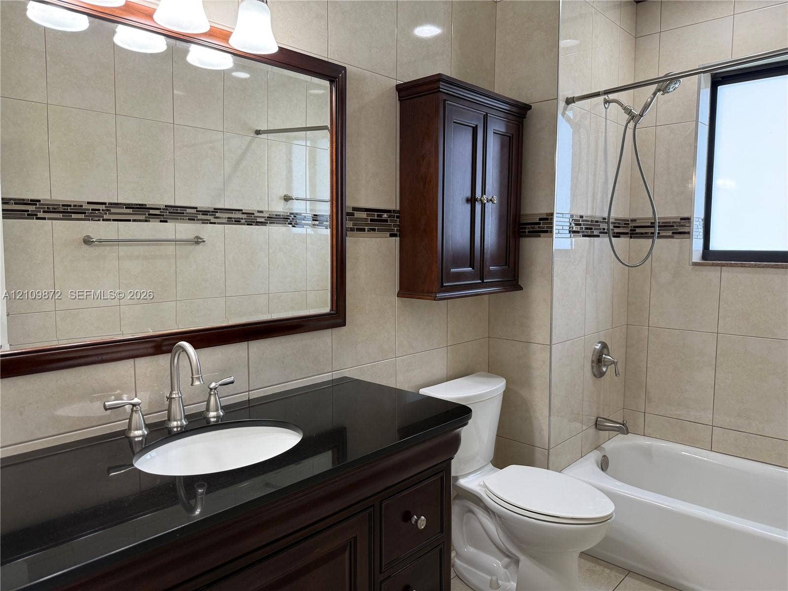 811 NE 10th Street, #5, Hallandale Beach, Hallandale Beach, FL 33009 Bathroom featuring a dark wood vanity with a black stone countertop, inset sink, full-width mirror with wood frame, coordinating wall-mounted storage cabinet, and floor-to-ceiling tile with decorative inlay