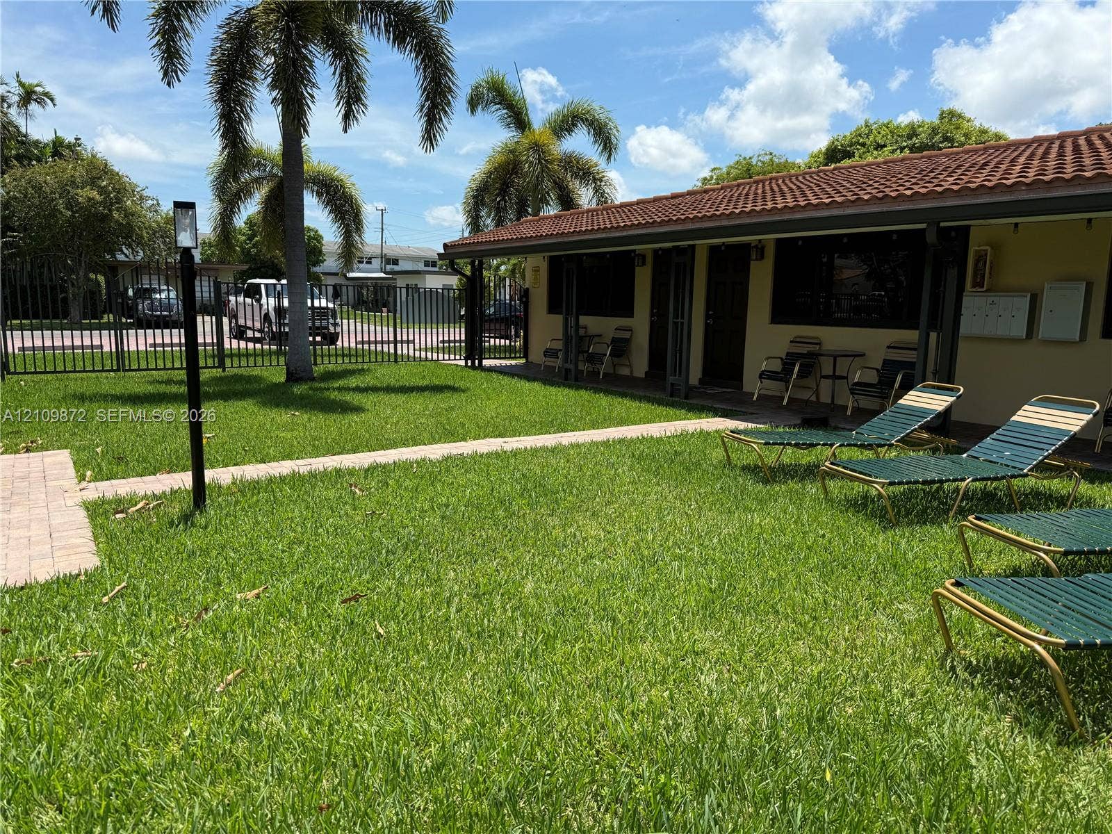 811 NE 10th Street, #5, Hallandale Beach, Hallandale Beach, FL 33009 Grassy outdoor space featuring a red-tiled roof, stone-paver walkway, metal perimeter fencing, and mature palm trees