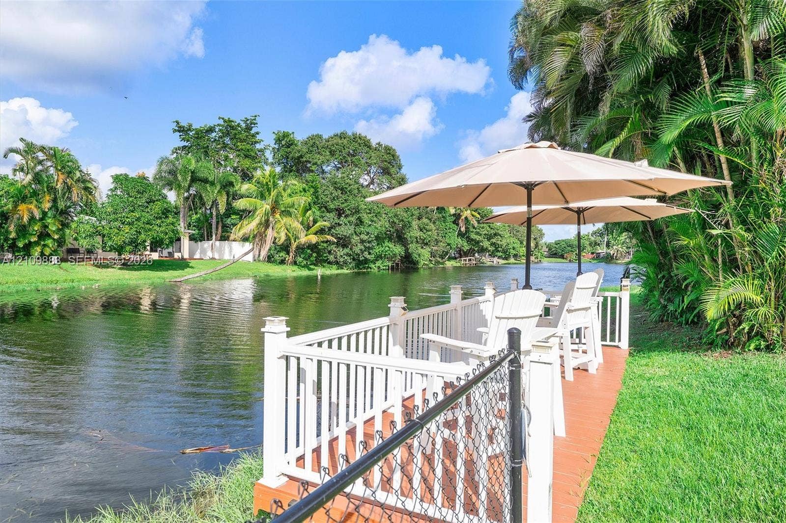 751 SW 54th Avenue, Margate, FL 33068 Waterfront deck featuring a wood-tone surface, white railing, and perimeter chain-link fencing, situated along a natural waterway with surrounding tropical landscaping