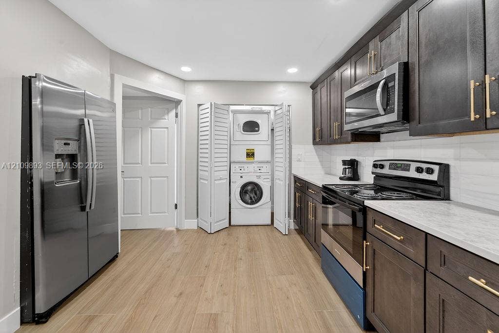 712 Tradewind Drive, North Palm Beach, North Palm Beach, FL 33408 Kitchen featuring dark-stained shaker cabinetry, stainless steel appliances, light-toned stone-finish countertops, wood-finish flooring, and an integrated laundry closet with bi-fold doors