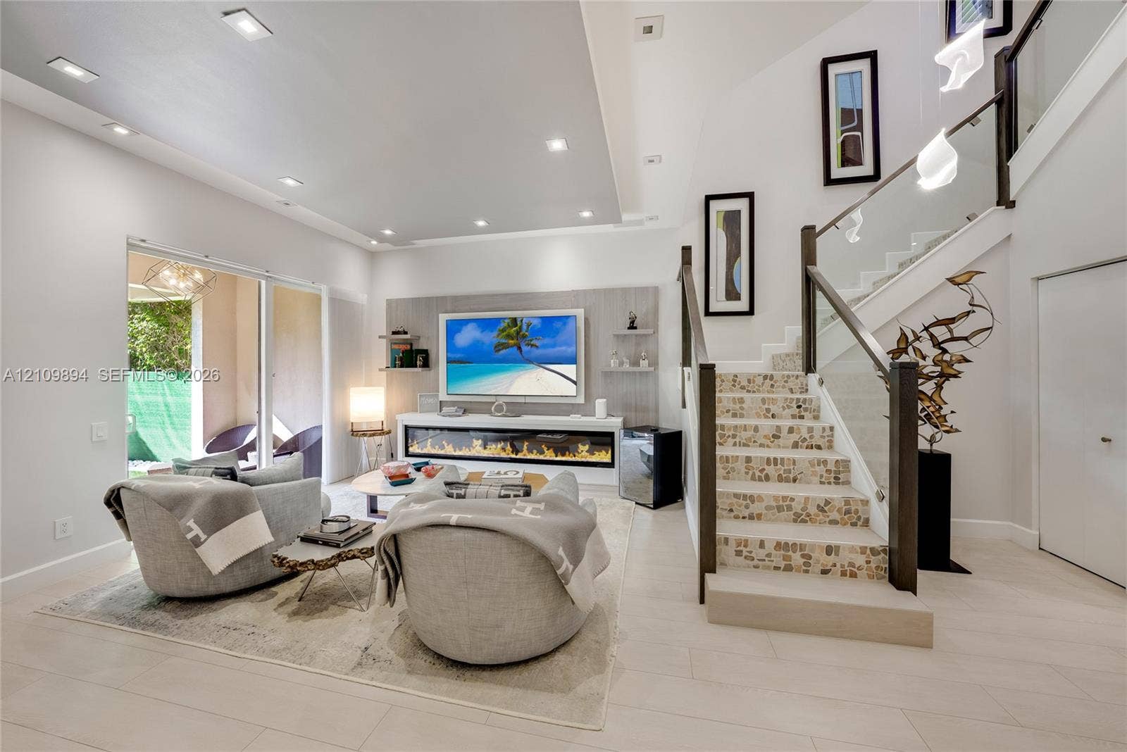 Open-concept living space featuring a recessed ceiling with integrated lighting, built-in linear fireplace with stone-finish surround, staircase with glass panel railing, and large sliding glass door