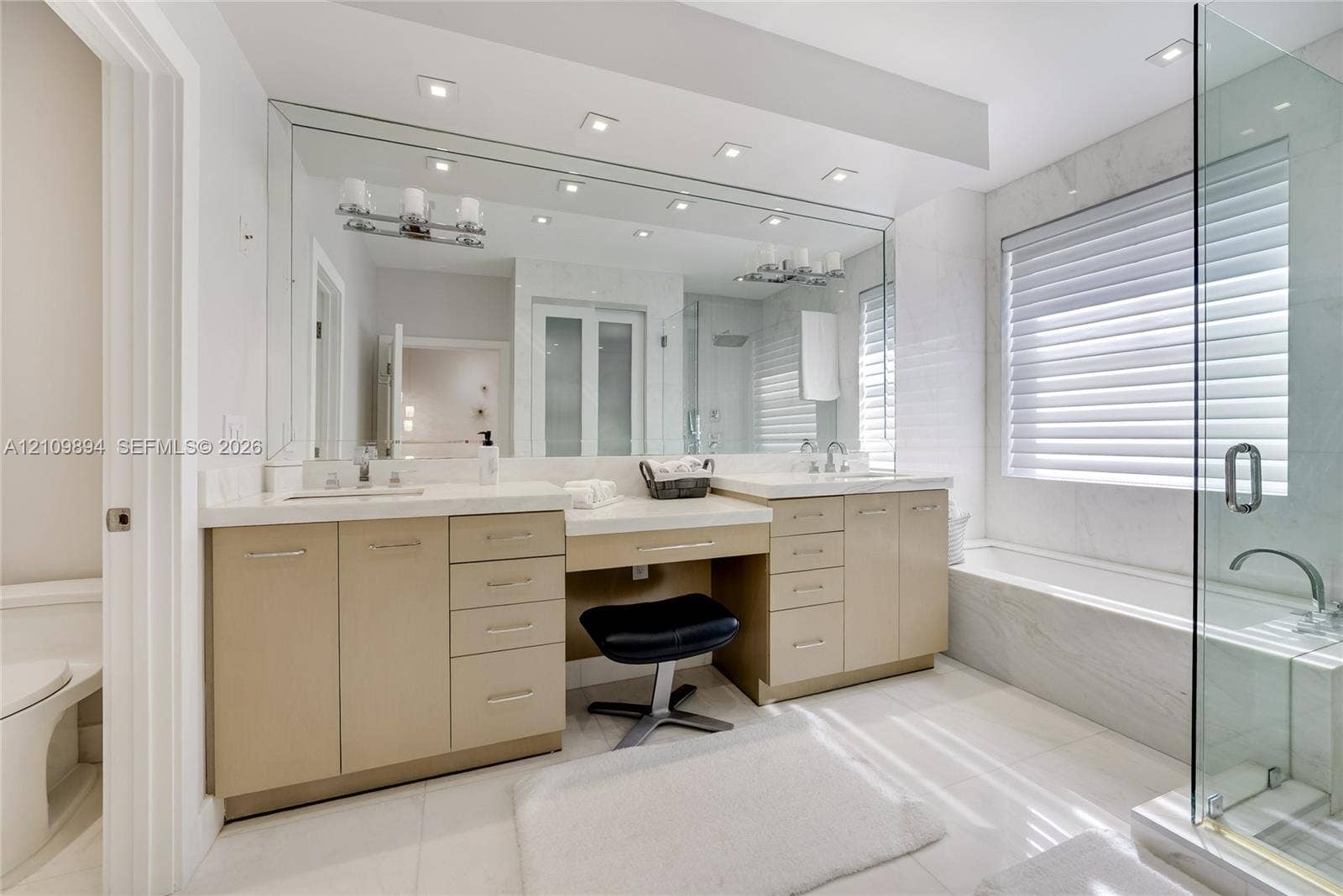 Double vanity featuring cabinetry with satin-finish hardware, integrated quartz countertops, an inset makeup station, full-width wall mirror, and a glass-enclosed shower with a deep soaking tub