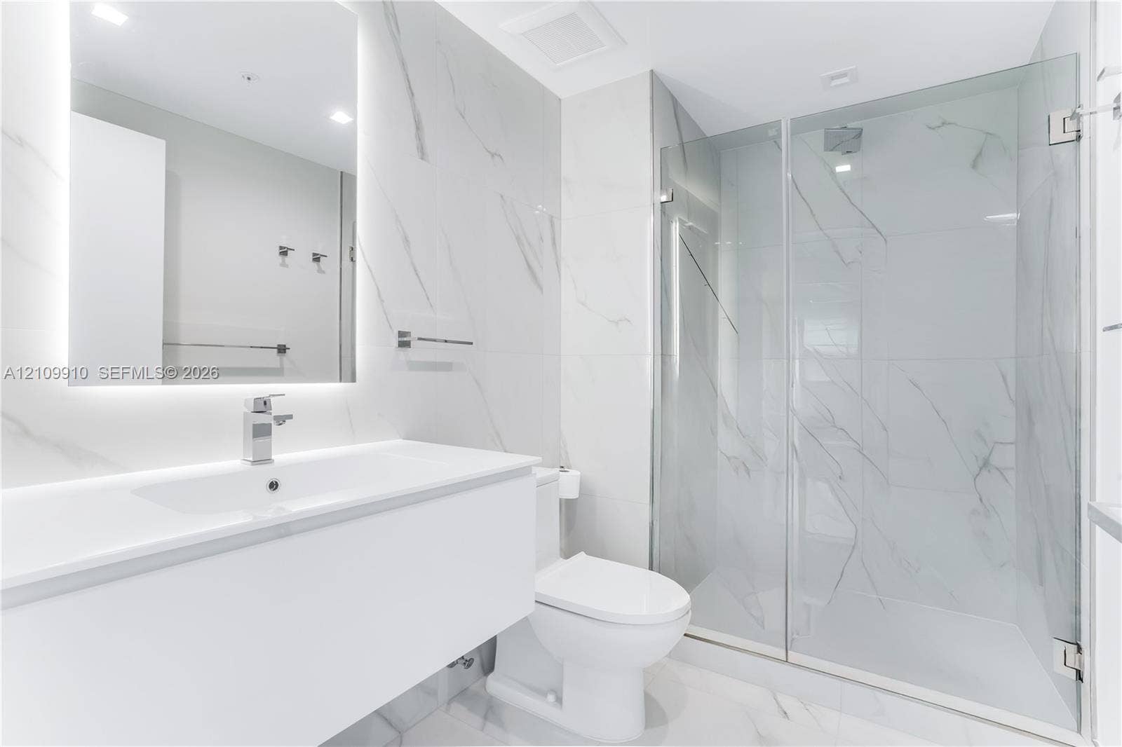 1170 E Hallandale Beach Boulevard, #908, Hallandale Beach, FL 33009 Bathroom featuring a floating vanity with a smooth surface, wall-to-wall mirror with integrated lighting, large-format stone-patterned wall tile, and a glass-enclosed shower