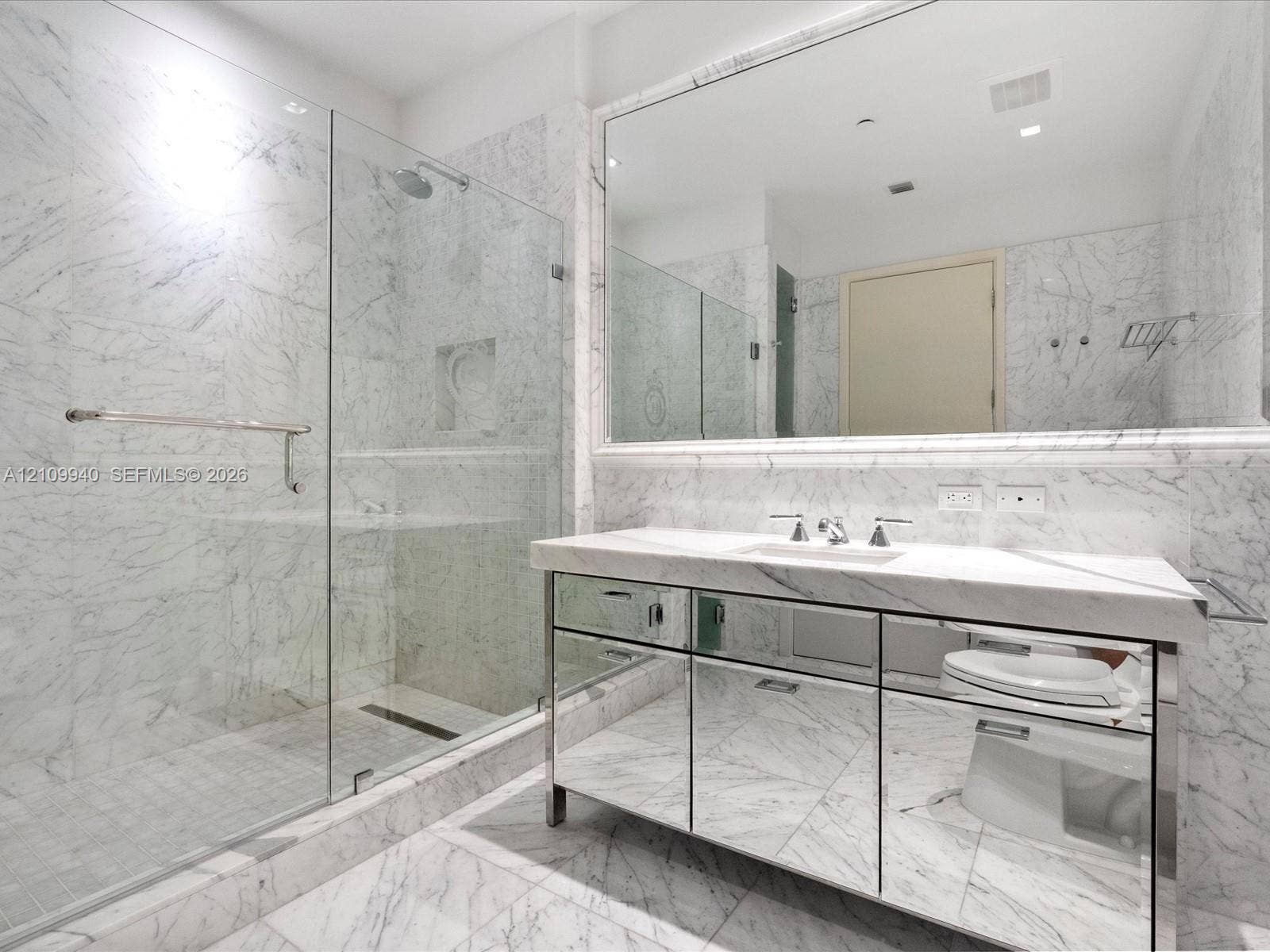 Full-wall stone tiling, glass-enclosed walk-in shower with recessed shelving, mirrored vanity cabinetry, stone-topped vanity, and expansive wall-mounted mirror