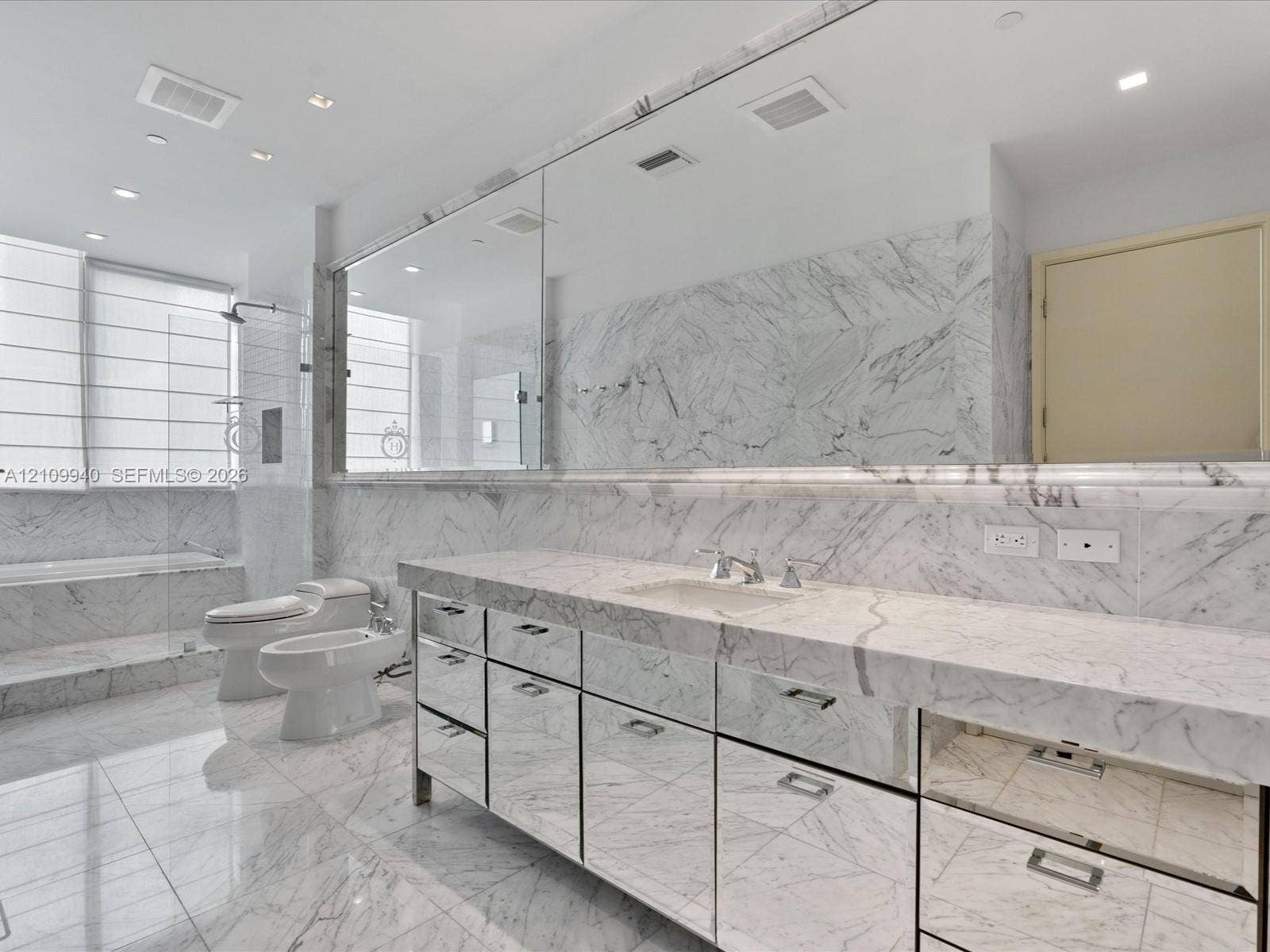Full-wall vanity mirror, custom cabinetry with mirrored drawer fronts, natural stone countertops and wall cladding, recessed lighting, and a separate glass-enclosed shower and soaking tub
