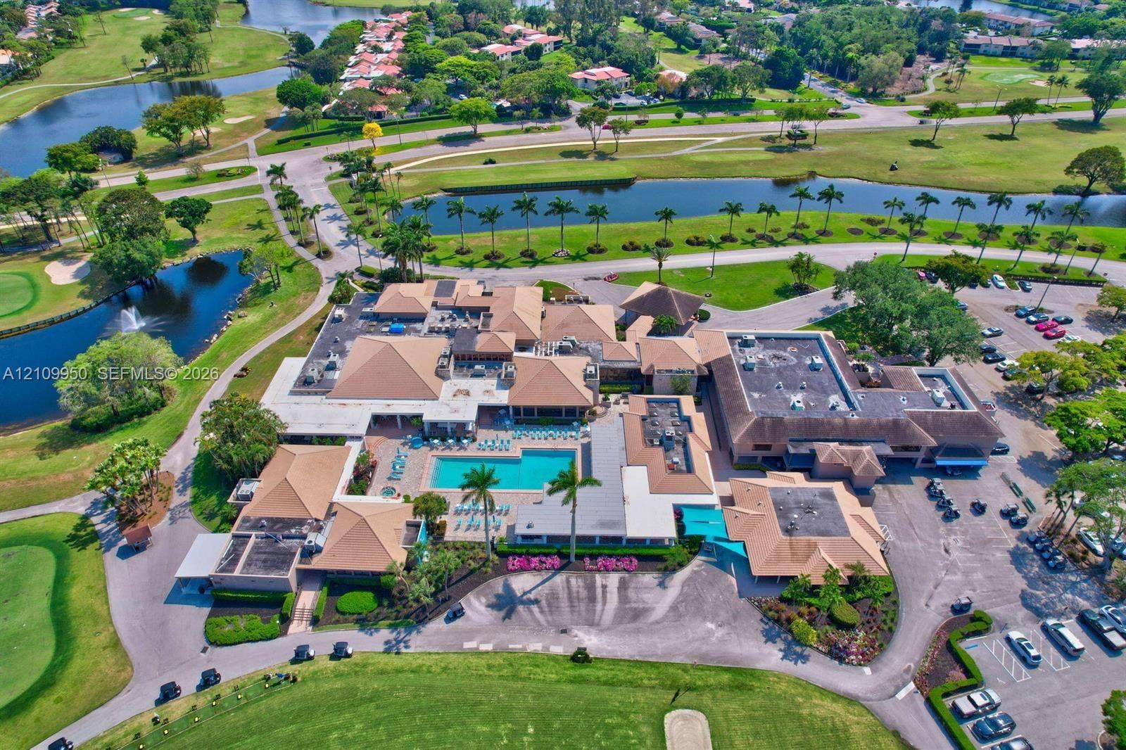 Aerial perspective of a multi-structure facility featuring a central swimming pool, paved surface parking, mature landscaping, and surrounding water features