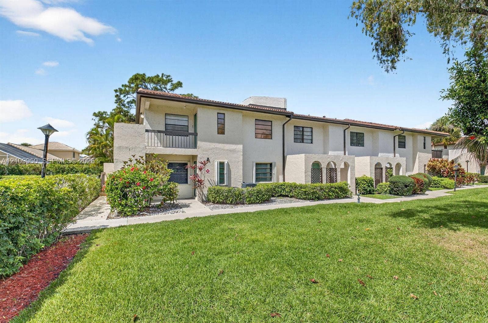 Multi-story stucco exterior featuring arched ground-level entryways, a second-story balcony with metal railings, tile roof, and concrete walkways
