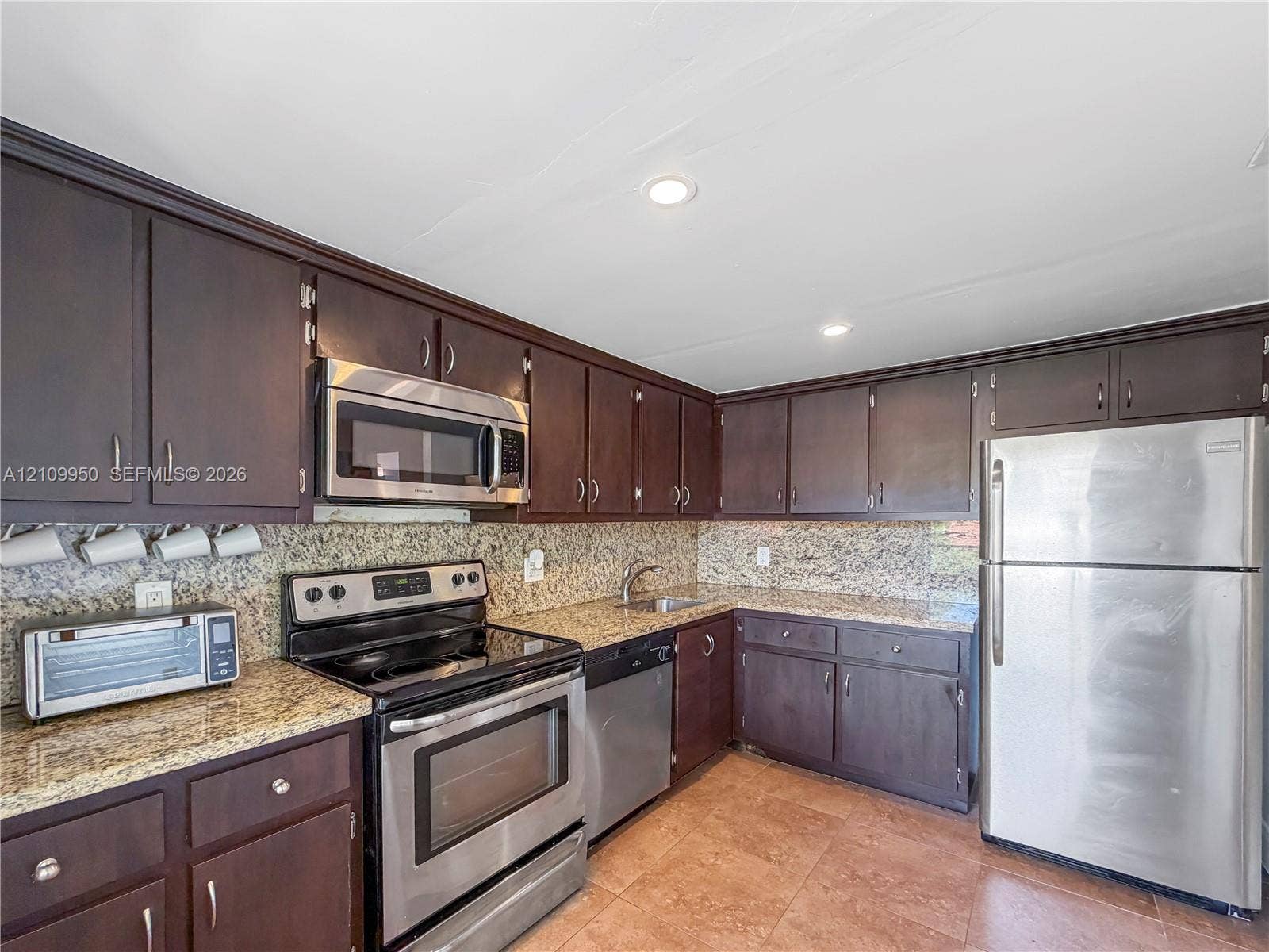 Kitchen featuring dark-finish wood cabinetry, speckled stone-finish countertops, tile flooring, and integrated stainless steel appliances