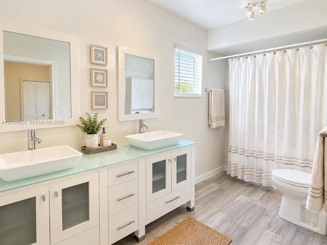 8528 Windsor Drive, Miramar, FL 33025 Bathroom featuring a dual-sink vanity with white cabinetry, frosted glass cabinet inserts, a light-toned countertop, rectangular vessel sinks, and wood-finish flooring