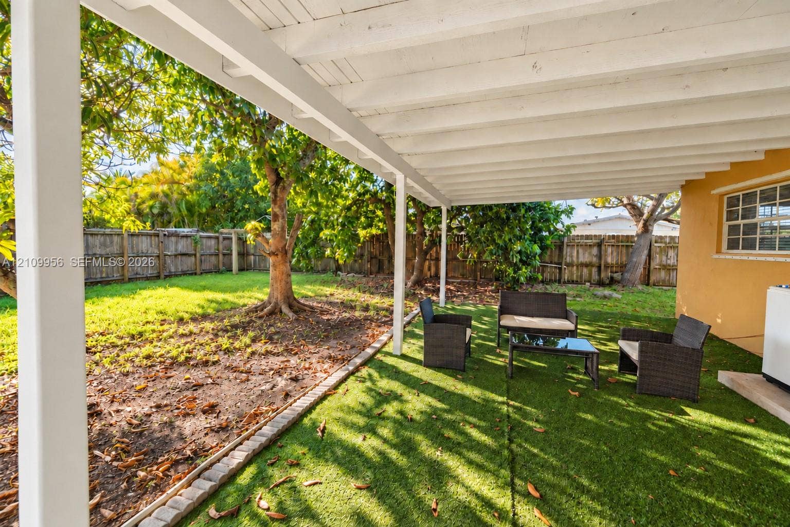 19503 SW 114th Place, Miami, FL 33157 Covered patio featuring a white exposed-beam ceiling and wooden support posts, complemented by a fenced backyard with artificial turf, stone border edging, and mature trees
