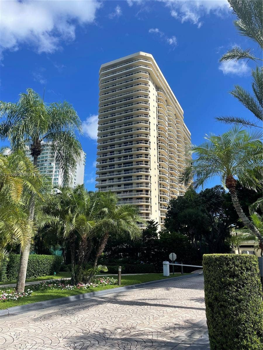 20191 E Country Club Drive, #2601, Aventura, FL 33180 High-rise residential building featuring stacked private balconies, a paved drive, manicured tropical landscaping, and mature palm trees