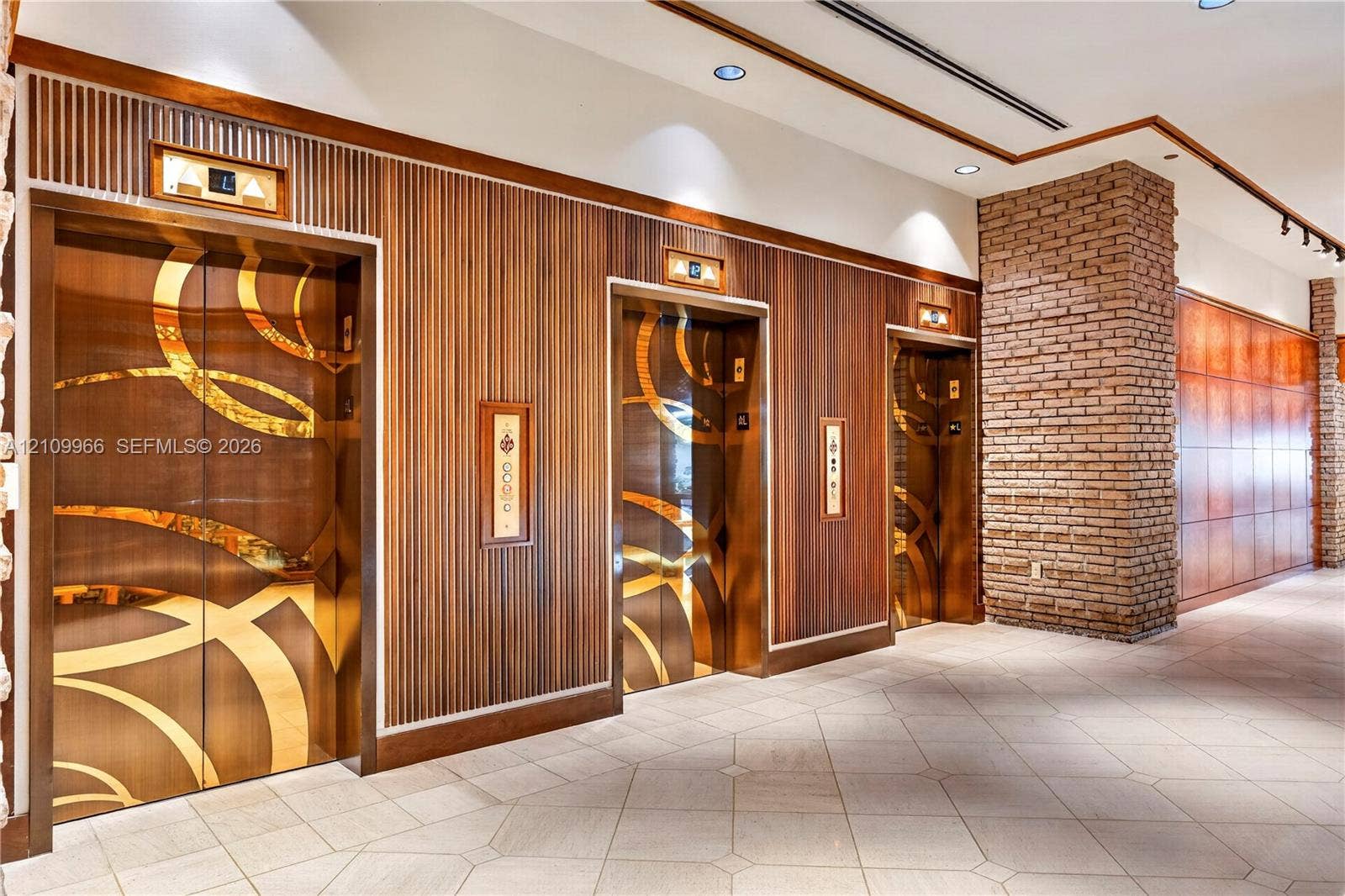 Elevator lobby featuring metallic geometric door panels, slatted wood wall cladding, exposed brick masonry pillar, recessed lighting, and tile flooring