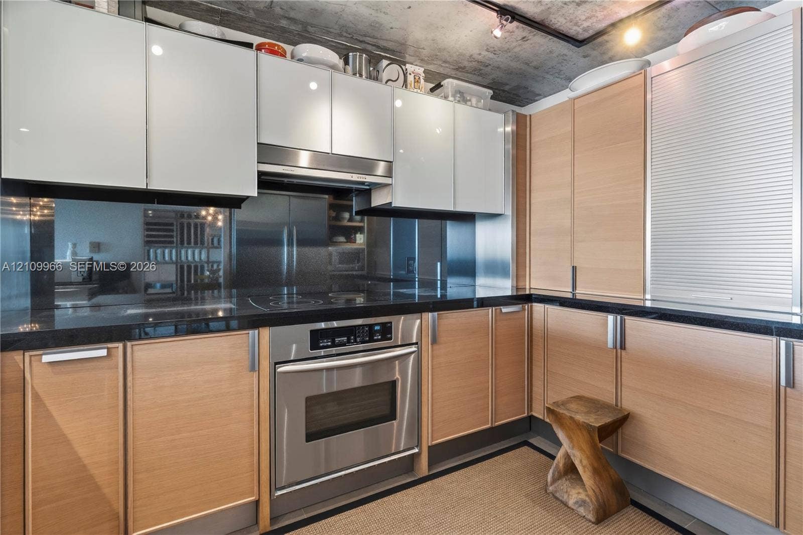 Kitchen featuring dual-toned flat-panel ceiling height cabinetry, dark stone countertops, integrated stainless steel oven, exposed concrete ceiling, and mirrored backsplash