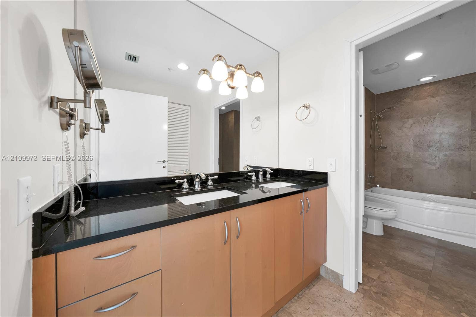 300 S Biscayne Boulevard, #T-1806, Miami, Miami, FL 33131 Dual-sink vanity featuring a dark stone countertop, light wood cabinetry, a large wall-to-wall mirror, and multi-light vanity lighting