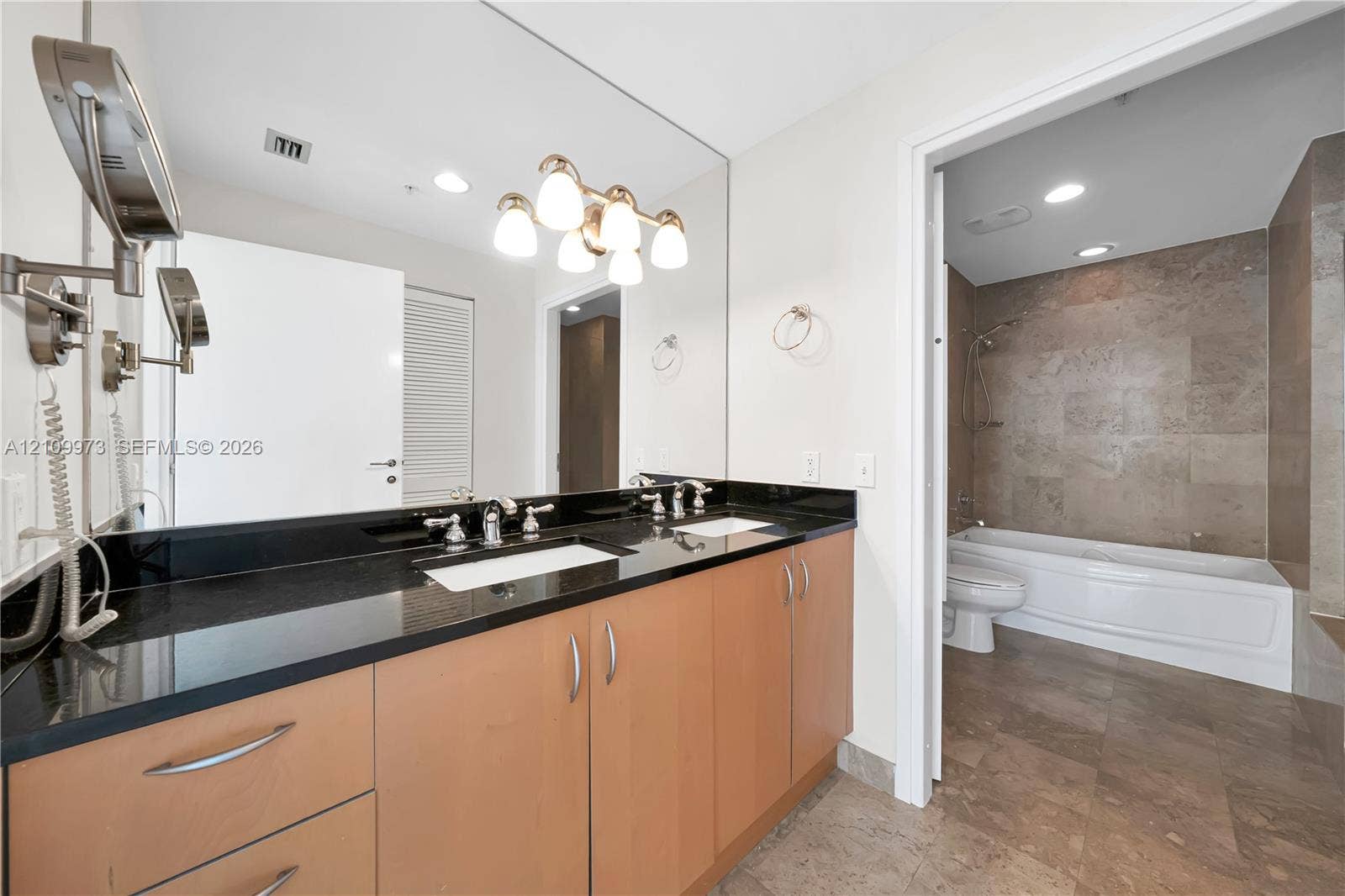 300 S Biscayne Boulevard, #T-1806, Miami, Miami, FL 33131 Dual-sink vanity with dark stone countertop, wood-finish cabinetry, full-width mirror, and multi-bulb vanity lighting, featuring a tile-surround bathtub