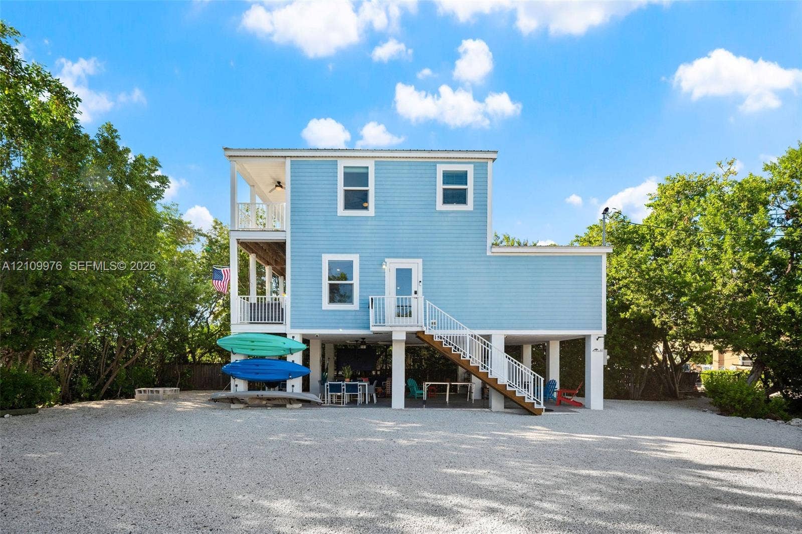 43 Waterways Drive, Key Largo, FL 33037 Elevated two-story residence featuring light blue lap siding, stacked covered porches, white metal railings, and a gravel ground-level parking area
