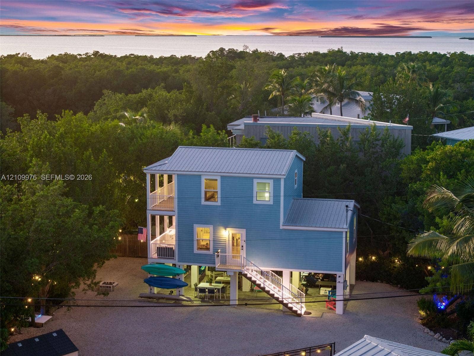43 Waterways Drive, Key Largo, FL 33037 Elevated coastal residence featuring a metal roof, multi-level covered porches, and a gravel ground-level space, set against a backdrop of mature foliage and open water