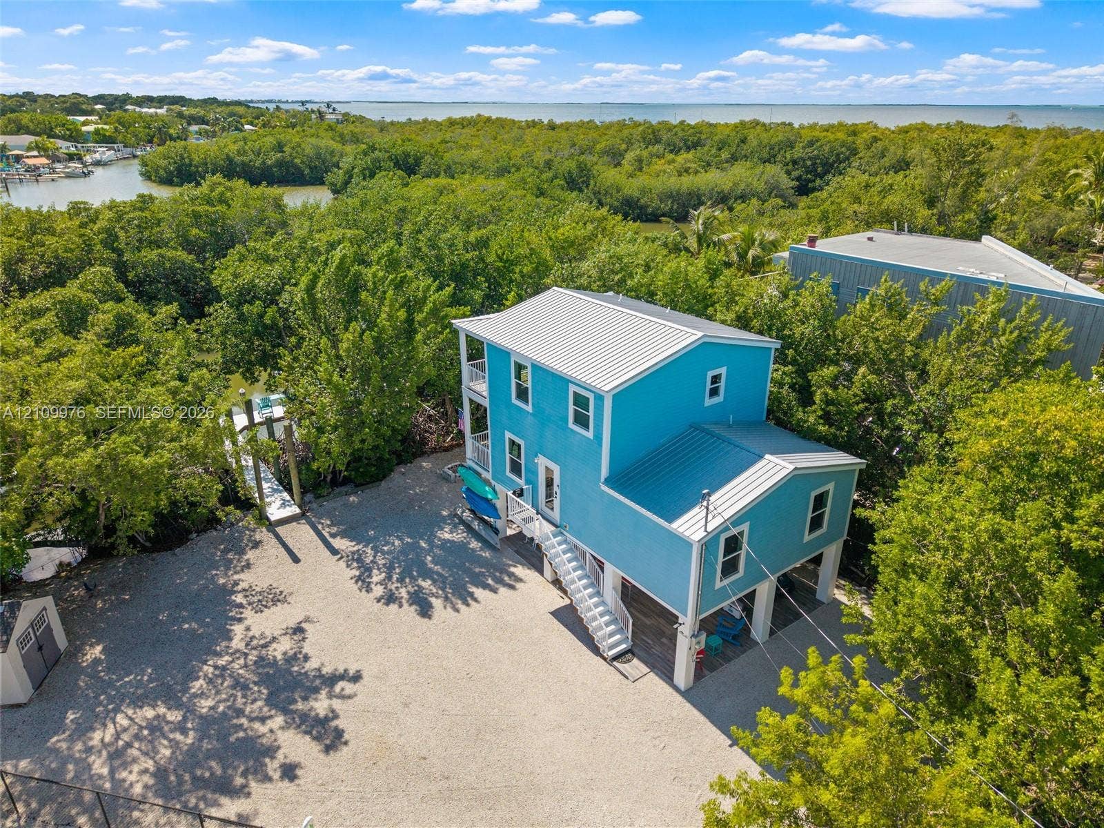 43 Waterways Drive, Key Largo, FL 33037 Two-story elevated coastal residence featuring a metal roof, wraparound exterior staircases, covered ground-level parking, gravel driveway, and a private dock