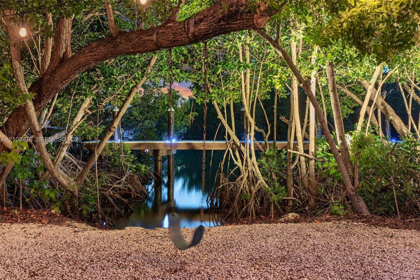 43 Waterways Drive, Key Largo, FL 33037 Waterfront grounds featuring a private wood dock, pebble-surfaced shoreline, mature coastal trees, and exterior string lighting