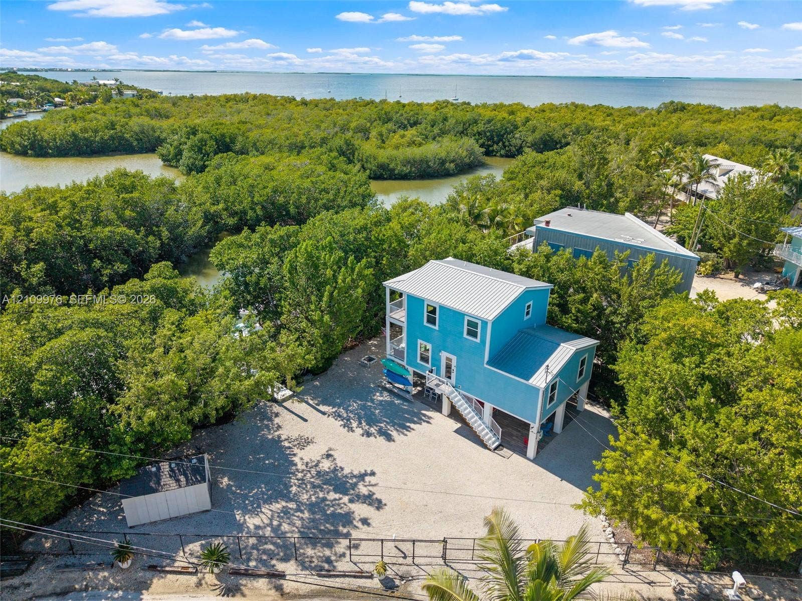 43 Waterways Drive, Key Largo, FL 33037 Elevated residence featuring a metal roof, exterior stairway, stone-surface driveway, and onsite storage structure, situated within proximity to coastal waterways and mangroves