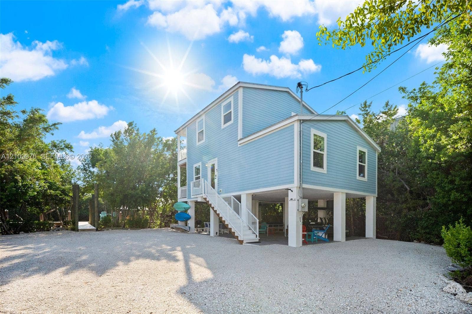 43 Waterways Drive, Key Largo, FL 33037 Elevated two-story residence with light blue horizontal siding, open-air ground level, exterior staircase with white railings, and gravel driveway