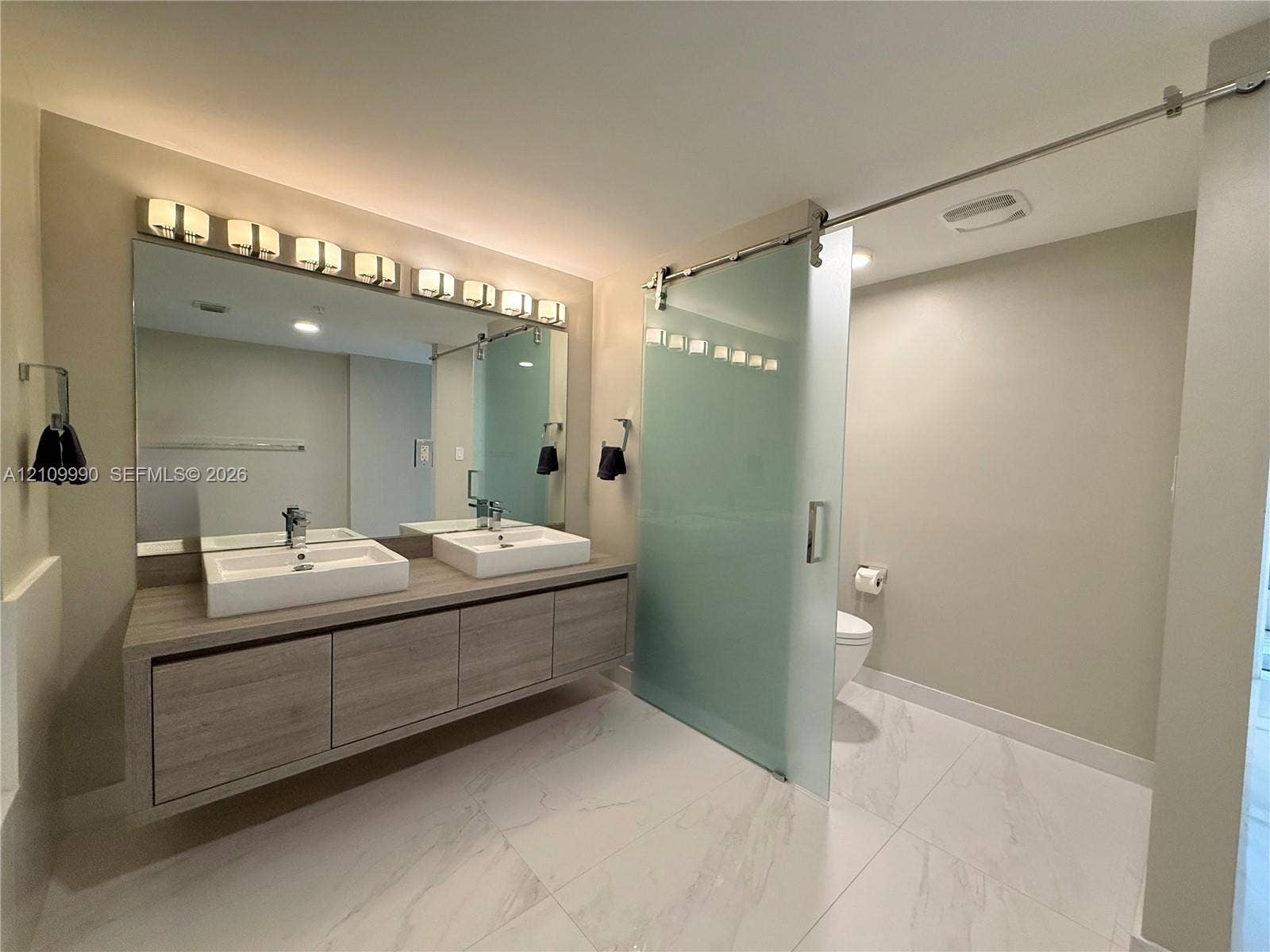 Floating wood-finish vanity with dual rectangular vessel sinks, expansive wall-to-wall mirror, oversized multi-bulb light fixture, and frosted glass sliding door