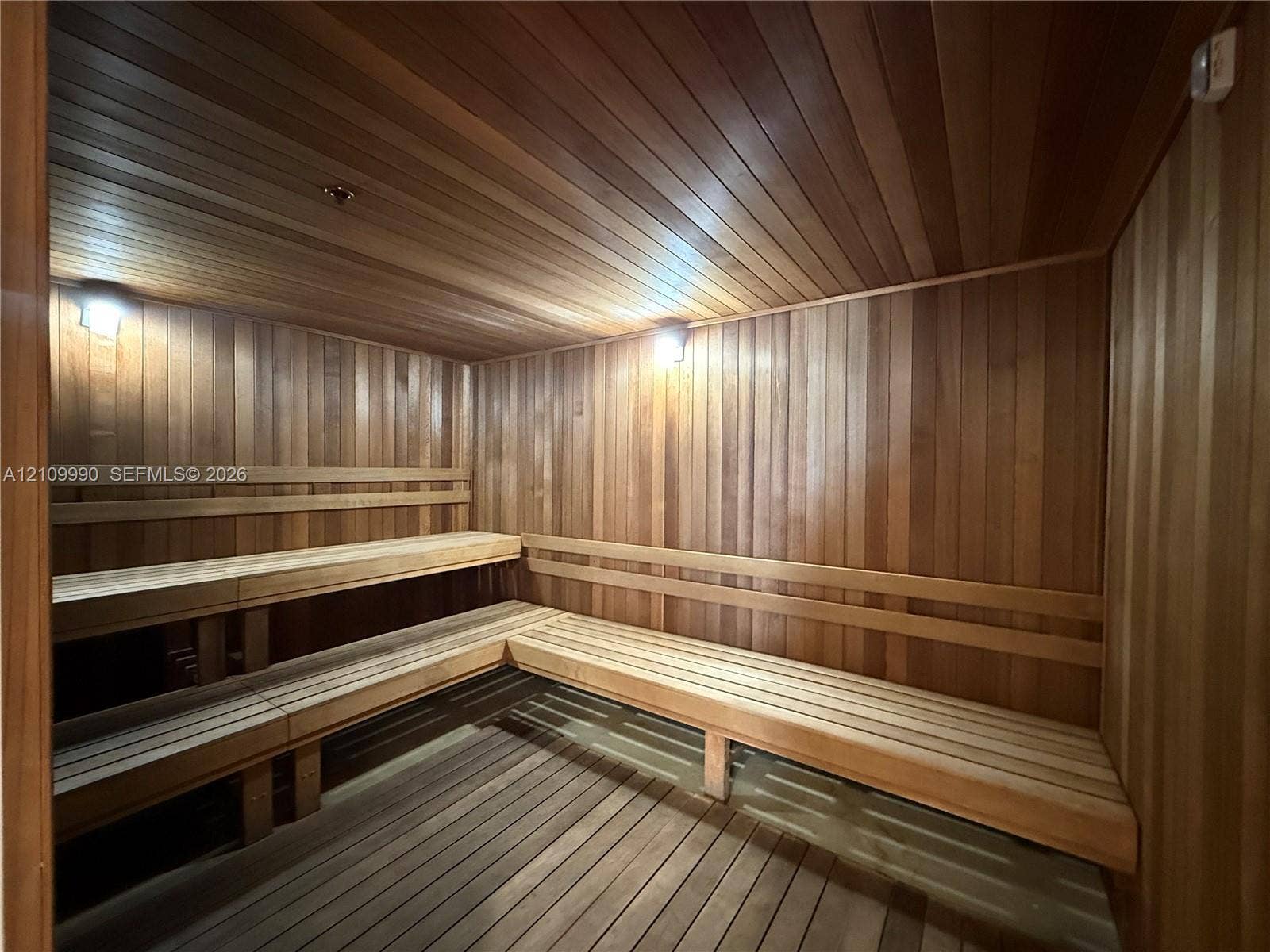Sauna featuring natural wood tongue-and-groove wall paneling, tiered bench seating, slatted floorboards, and wall-mounted lighting