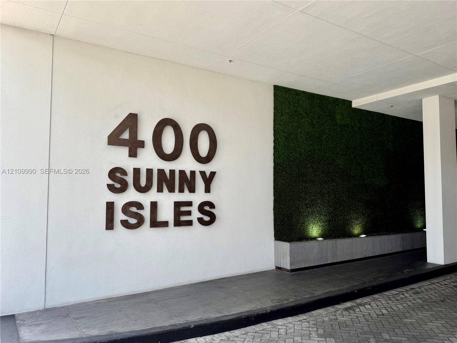 Covered entryway featuring dimensional signage, a green wall accent, concrete planter base, and stone paver driveway