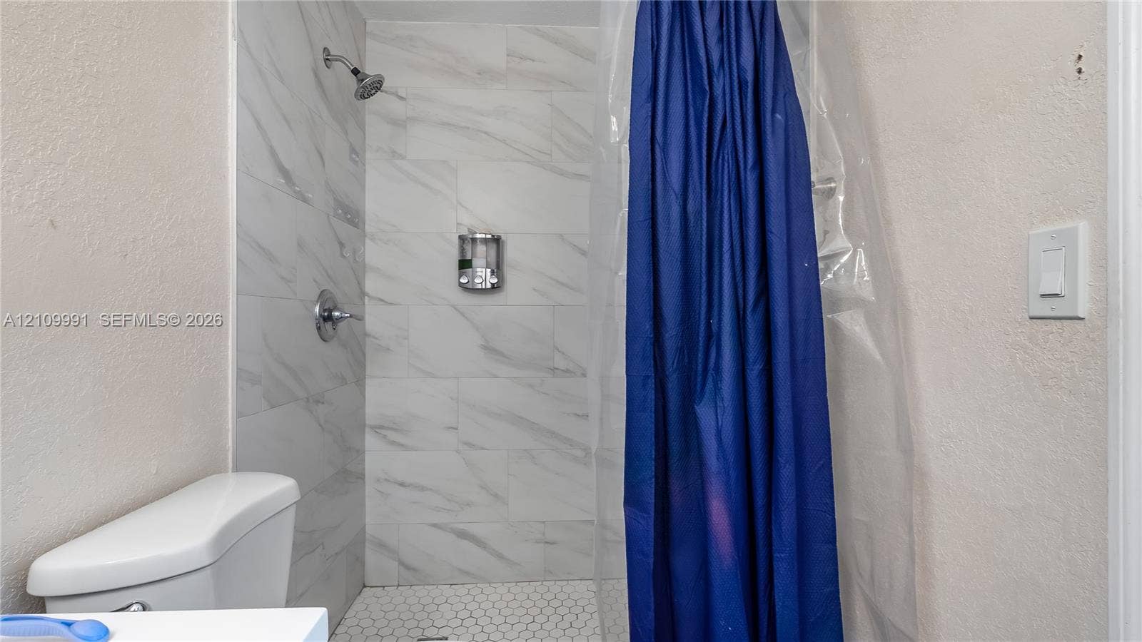 Shower stall featuring large-format wall tile with a veined pattern, hexagonal floor tile, chrome shower fixture, and wall-mounted dispenser