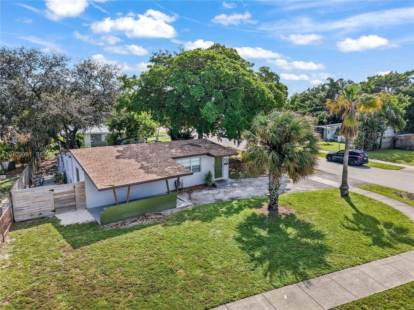Single-story exterior featuring a shingled roof, overhanging carport, integrated wood-finish fencing, and established palm and shade trees
