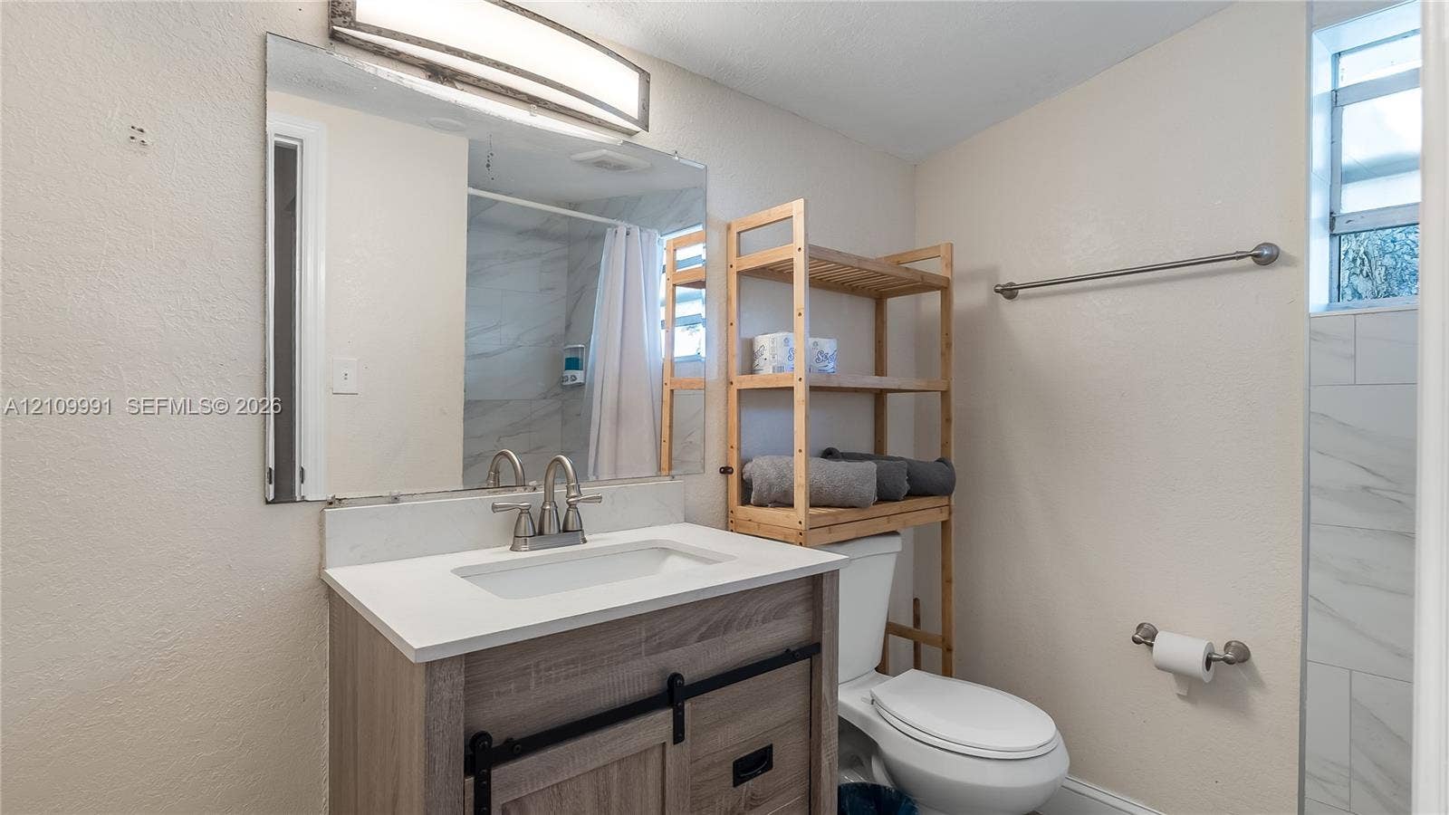 Bathroom featuring a wood-finish vanity with a stone-composite countertop, undermount sink, dual-handle faucet, large wall-mounted mirror with integrated lighting, and a shower area with light-toned tiled walls