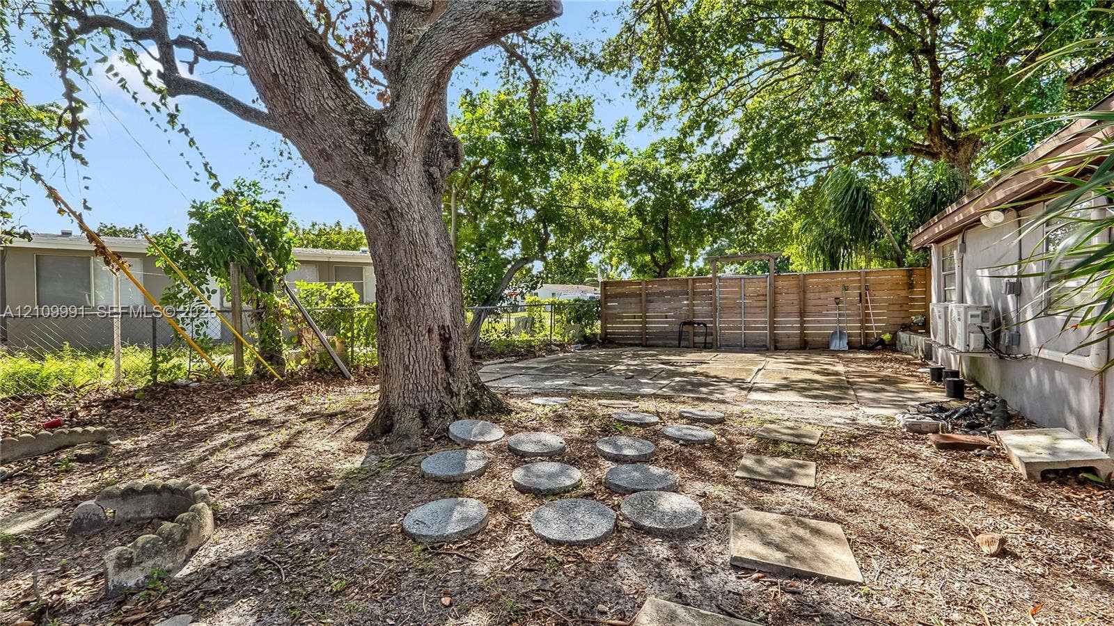 Rear exterior area featuring a concrete patio, mature tree, wood-slat privacy fencing, and concrete stepping stones
